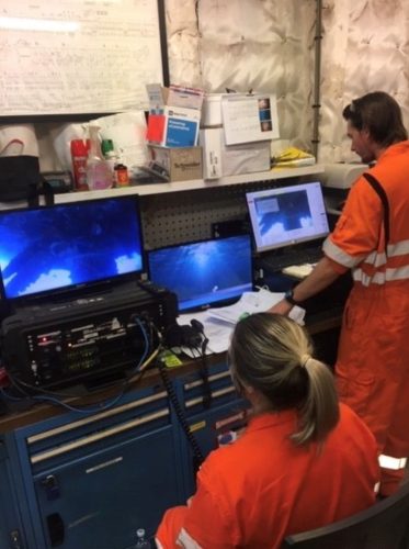 Two technicians in high-visibility orange workwear are operating sophisticated electronic equipment in an industrial environment. One technician is seated in front of multiple monitors displaying underwater ROV (Remotely Operated Vehicle) operations, while the other, standing, examines data or controls on another screen. Their workstation is cluttered with various operational manuals, supplies, and personal items, indicating a busy and dynamic work setting. The focus and teamwork between the individuals suggest a critical monitoring or controlling task.
