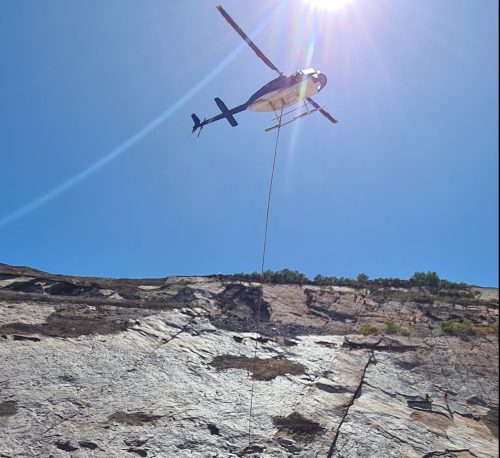 An underside shot of a helicopter lowering a roll of TECCO mesh on rope to two IRATA Technicians on a rock face.