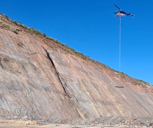 A helicopter lowering a roll of TECCO Mesh on rope to rope access technicians to apply to the rock face.