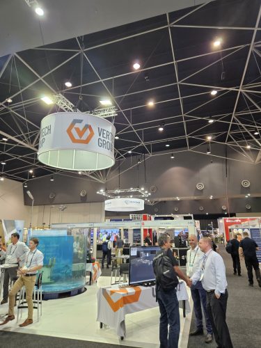 The Vertech Group's AOG Energy 2023 exhibition stand is prominent inside a busy trade show hall, with a large circular hanging banner displaying its logo. Below, a clear cylindrical tank simulating a marine environment catches the eye, with attendees around, including three men, in a handshake and conversation. Overhead, the geometric ceiling pattern formed by interconnected metal beams and lighting fixtures gives an industrial feel to the scene, while various other exhibition booths and visitors in the background add to the event's bustling atmosphere.