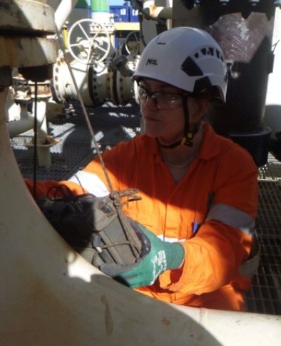 A worker wearing high-visibility orange coveralls and a white safety helmet inspects equipment on an industrial site. The worker wears safety glasses, ear protection, and green gloves and is focused on adjusting or examining a part of the machinery with their gloved hands. The background is occupied by industrial pipework and valves, suggesting a setting within a facility such as a plant or refinery. The worker's attention to detail underscores the importance of safety and precision in such an environment.
