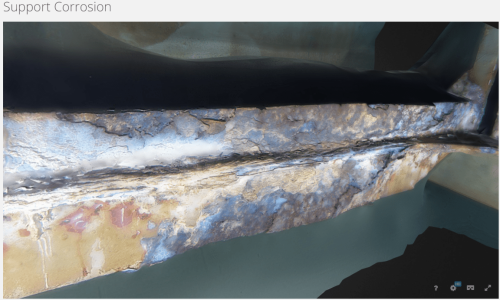 The image displays a 3D render of a structural support exhibiting significant corrosion. The metal surface is patchy with flaking rust and discolouration areas, ranging from dark grey to brown, and hints of the original paint in places. The visible deterioration suggests material degradation due to environmental factors. The text "Support Corrosion" indicates that the purpose of the image is likely for assessment and documentation of the damage to the structure's integrity. The background is out of focus, emphasizing the corrosion on the metal element in the foreground.