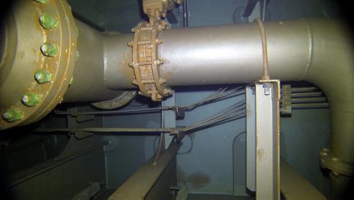 This image was taken within a ship's ballast tank, with a prominent thick pipe running horizontally across the frame. It's fitted with large bolted flanges, indicative of high-pressure capacity. Auxiliary pipes and metal supports surround the main pipe, while cables and conduits are neatly organised along the wall in the background. The scene is illuminated by artificial lighting, casting a greenish hue over the metal surfaces, which are coated with paint or protective layers showing signs of wear. The overall setting suggests an environment focused on utility and function, which are characteristics of a facility like a ship's interior or industrial plant.