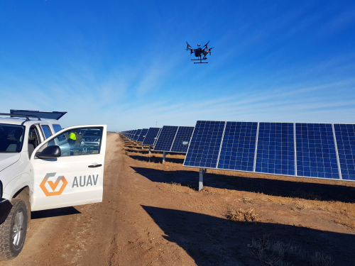 A drone hovers over a solar farm in a clear blue sky, with rows of photovoltaic panels stretching into the distance. On the left, a vehicle with the logo "AUAV" is parked, its door open, and a person in high-visibility clothing can be seen inside, suggesting they are operating the drone. The scene captures a moment of renewable energy technology being monitored or surveyed with advanced unmanned aerial vehicle equipment, highlighting innovation in sustainable energy management.