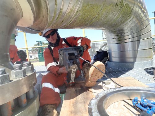 A technician sits under a bend pipe controlling the unseen olympus digital camera via a cord connected to an electronic device he is holding.