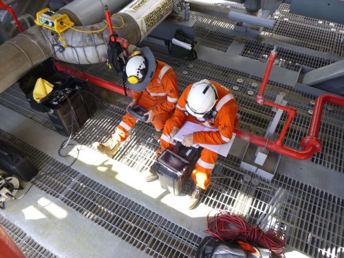 Two technicians are sitting on a pipe above a floor of metal grating; one is operating an electronic device, and the other is writing in a notebook.