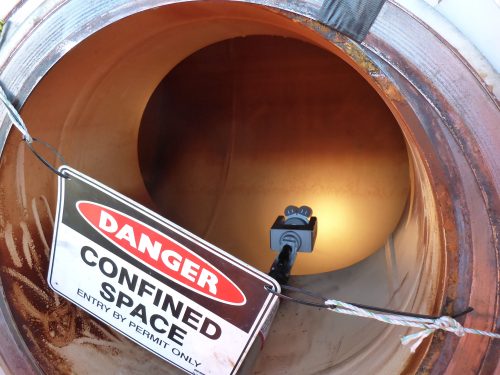 The entrance to a pipe is restricted by a sign reading "Danger: Confined Space"; beyond it, the Olympus digital camera is being inserted.