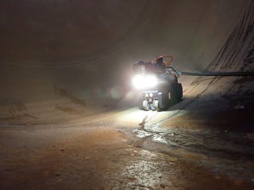 An RDVI robot with a front light on is driving along the floor of a pipe.