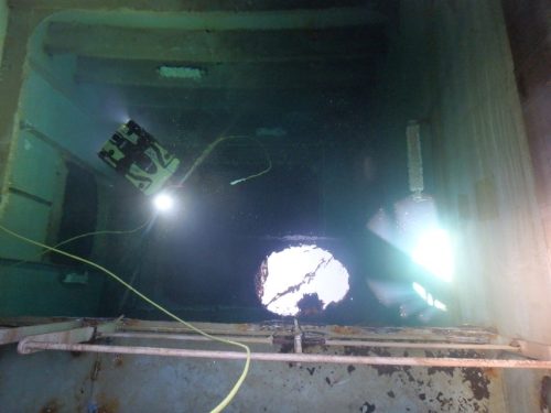 The image shows a dimly lit industrial interior viewed through a round hatch opening. Inside, we see a remotely operated vehicle (ROV) with bright lights and a patterned design suspended in water, indicating underwater operations, possibly for inspection or repair. Yellow cables connected to the ROV extend out of the hatch, contrasting with the structure's dark waters and rusted metal surfaces. A beam of light penetrates the water from the ROV's lamp, illuminating the murky surroundings. The scene captures a moment of technical marine operation, highlighting the interaction of technology and the challenging aquatic environment.