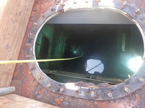 The image shows a close-up view of a round metal hatch with a heavily rusted and bolted flange on a structure that appears industrial, possibly part of a vessel or tank. The dimly lit interior is partially visible through the hatch, featuring more rust and metal framework, indicating an aging or long-used facility. A yellow cable extends through the hatch to an ROV, suggesting ongoing work or inspection activity. The photograph captures the weathered textures of the metal and the contrast between the dark inner environment and the brighter exterior.