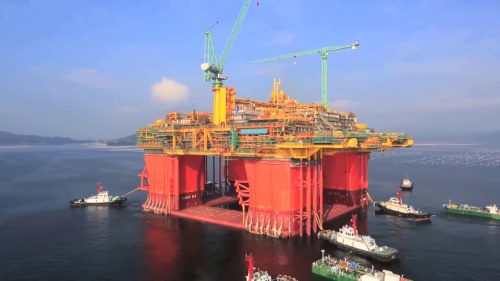 A massive offshore oil platform stands majestically in calm sea waters, painted in bright red and yellow. The platform has heavy industrial equipment, including cranes and drilling infrastructure. Several support vessels, including tugs and supply boats, are alongside the platform, indicating active operations. In the background, a serene coastline stretches across the horizon beneath a clear blue sky. The ocean reflects the sunlight, creating a shimmering effect on the water's surface.