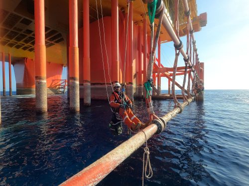A Vertech IRATA Rope Access Technician hangs suspended from the side of an FPSO with ropes, the ocean visible in the background.