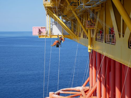 A vertech IRATA Rope Access Technician hangs suspended from the side of a fpso with ropes, the ocean visible in the background.