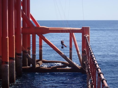 A vertech IRATA Rope Access Technician hangs suspended from the side of an FPSO with ropes, the ocean visible in the background.