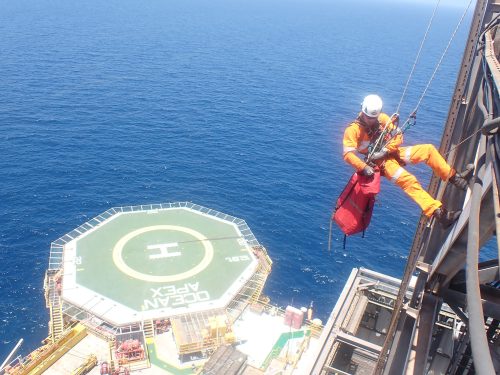 A vertech IRATA Rope Technician descends off the side of a derrick with a helicopter landing pad and ocean in the background.