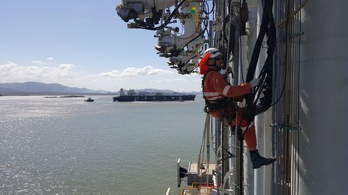 A vertech IRATA Rope Access Technician in the process of scaling the side of the Gladstone LNG Facility - ocean and a barge visible in the background.