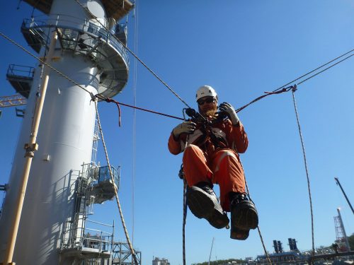 A Vertech IRATA Rope Access Technician poses for a photo while suspended by multiple ropes on the Gladstone LNG Facility.loading=