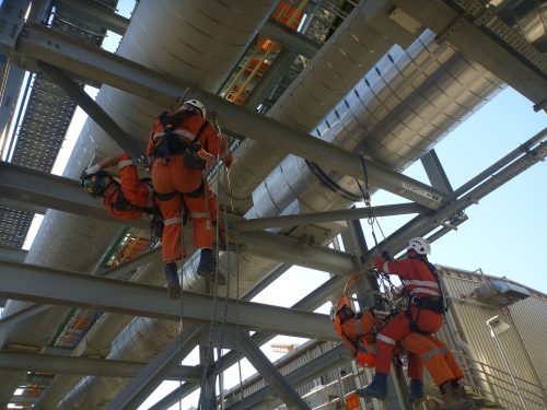 Four technicians in orange high-visibility workwear and safety harnesses are working on a large industrial pipeline as part of a shutdown service at the Gladstone LNG Facility. They are suspended above the ground by safety ropes, using hand tools. The pipeline runs alongside a complex metal framework, indicative of a substantial industrial or energy facility. The image focuses on the workers carrying out maintenance or inspection tasks on the pipeline's exterior.