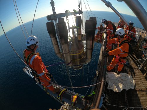Workers in high-visibility orange safety gear are conducting a flare tip changeover, with a clear sky and calm sea in the background. They are focused on maneuvering the old flare, which is being hoisted by a crane. One worker is standing on a platform overseeing the operation, secured with safety harnesses. The photo captures offshore industrial work's dynamic and potentially hazardous environment, emphasising safety and teamwork.