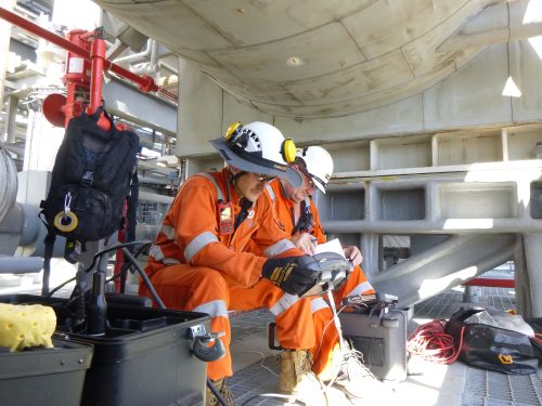 Two vertech inspectors sit on a raised platform under a large pipe using electronic equipment to monitor the testing process.