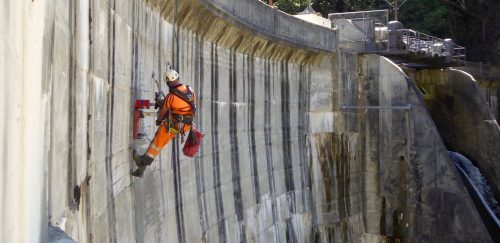 A worker in bright orange high-visibility clothing is abseiling down a large concrete dam wall. The worker is equipped with a safety harness, helmet, and various tools, indicating that they are performing an inspection or maintenance task. The scale of the person against it emphasises the sheer size of the dam wall. In the background, the top of the dam and the surrounding environment can be seen, highlighting the challenging and vast nature of the structure that requires such vertical work.