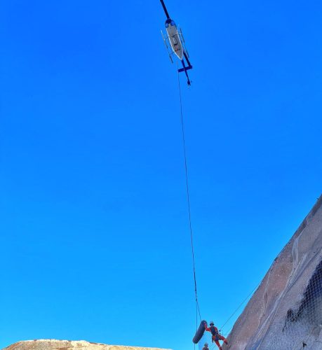 A worker in orange and white protective gear is suspended from a helicopter by a cable. The worker appears to be conducting an aerial operation or inspection on a large, steep slope reinforced with netting, likely for stabilisation. The backdrop is a clear blue sky, indicating fair weather, essential for helicopter operations. The high contrast between the vivid blue sky and the worker's outfit highlights the operation's aerial nature and the worker's safety gear.