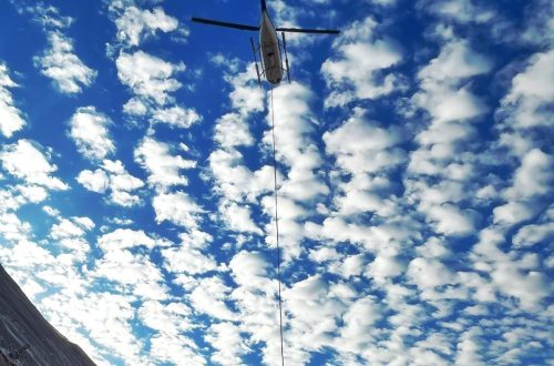 A helicopter hovers in a vibrant blue sky, scattered with fluffy white clouds. A cable extends from the helicopter to the bottom left of the frame, suggesting a winching operation. The corner of a steep grey cliff is visible, hinting at the challenging environment in which the helicopter operates. The perspective is upward-looking, capturing the grandeur of the sky and the meticulous nature of aerial work.