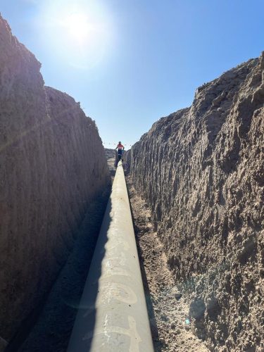 A trench dug into the dry, earthen ground under a clear blue sky. A single, long pipeline is installed within the trench, running straight through the centre of the frame. Sunlight floods the scene from the top left corner, causing a slight flare in the image and highlighting the textures of the dirt walls. A figure is visible at the front end of the trench, standing and looking towards the camera, their shadow stretching long beside the pipeline. The depth of the trench about the standing figure suggests it is several feet deep.