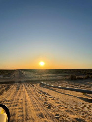 The sun sits low in the sky, casting a warm golden glow over a sandy landscape with tyre tracks marking the path. A pipeline runs parallel to the path on the right-hand side, partially buried in the sand. Sparse vegetation is visible in the distance, and safety markers are positioned at intervals alongside the pipeline. The shadow of the vehicle from which the photo was taken stretches towards the sunset, suggesting the image was captured during early morning or late afternoon.