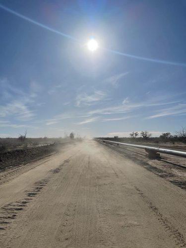 A bright sun hangs high in a hazy blue sky, casting a soft glow over a dusty dirt road. The road stretches straight into the horizon, flanked on the right by a long pipeline supported by stands above the ground. The glare from the sun creates a slight lens flare, and thin clouds are barely visible in the sky. The scene is tranquil, with no visible movement except the swirling dust, which suggests a breeze across the otherwise still landscape.