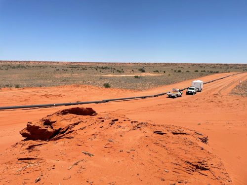 Two vehicles, a four-wheel drive and a ute, are stationary beside a long pipeline on a vast red sandy terrain. The pipeline extends across the image, disappearing into the horizon under a cloudless sky. The terrain is flat with sparse green shrubbery, typical of arid or desert regions. The bright sunlight casts short shadows from the vehicles, indicating a time around midday. The environment is reminiscent of a remote industrial site in a desert setting.