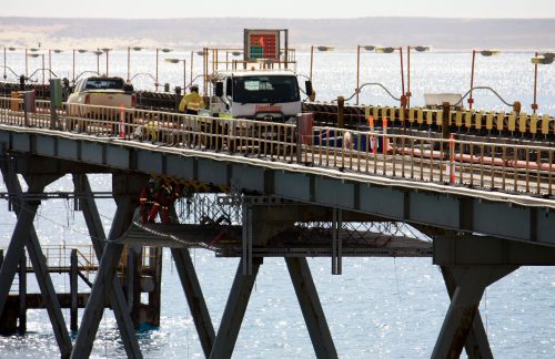A vertech technician in yellow fluro stands next to a flatbed truck on a bridge over water; decking can be seen on the bridge's underside.