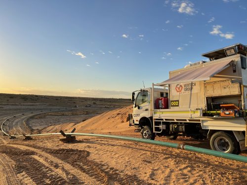 A mobile workspace mounted on the back of a white truck, labelled "Vertech Group," is stationed on sandy terrain with sparse vegetation under a clear, early evening sky. A pale green pipe runs parallel to the truck, and tyre tracks are imprinted in the sand, suggesting recent activity. The setting sun casts a warm glow on the scene, highlighting the contours of the sandy landscape. The open side panel of the truck reveals equipment and supplies, indicating an operational work site.