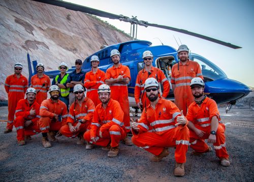 A group of IRATA Rope Access Technicians posing for a group photo in front of a helicopter used to deliver meshing to the rock face.