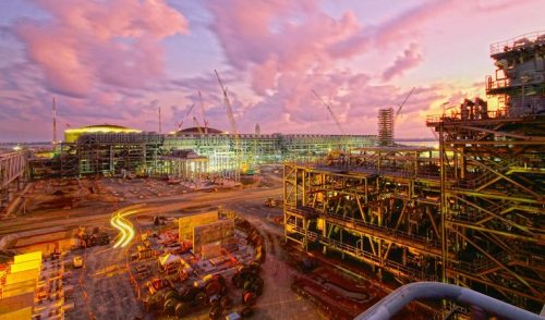 Twilight descends over the construction of the INPEX Ichthys facility. Towering cranes and scaffolding silhouettes against the dramatic sky, painted with orange, pink, and purple streaks from the setting sun. A labyrinth of steel structures reveals the complexity of the facility, highlighted by the warm glow of artificial lights. Foreground activity shows construction vehicles and equipment scattered throughout the site, indicating ongoing development. The scene captures the energy of industrial growth set against the backdrop of a vivid sky at dusk.