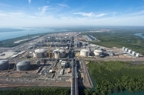 An aerial view of the INPEX ILNG Facility. The site is densely packed with storage tanks, piping, and steel structures, indicating a possible oil refinery or chemical plant. Large cylindrical tanks are prominent, with some under construction, evidenced by cranes and scaffolding. A natural water body borders the complex on one side and a green forested area on the other, contrasting industrial activity and natural landscapes. Roads and vehicles are visible, and a jetty extends into the calm blue waters, where a ship is docked. The sky is clear with a few scattered clouds.