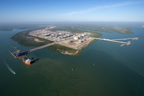 An aerial view of the INEPX LNG facility in Darwin, with multiple storage tanks and structures. A long jetty extends into the calm blue waters, with a large cargo ship docked at one end and a smaller vessel nearby, creating a wake. The surrounding area is lush with greenery, and the horizon shows a vast expanse of mangroves fading into the distance. The sky is clear, and the water reflects the sky's blue, hinting at an ecosystem coexisting alongside industrial activity.