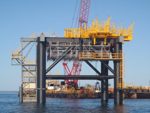 A view from the ocean of an offshore platform with a barge behind it.
