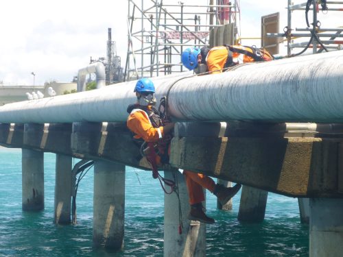 A vertech technician suspended from a supporting beam on an industrial jetty.