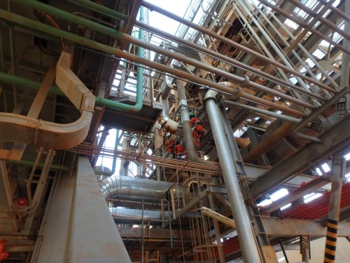 A complex array of industrial pipes and structures within a large facility. The image shows an intricate network of piping, both horizontal and vertical, with some wrapped in insulation. There's scaffolding suggesting maintenance or construction work, and a few workers wearing safety harnesses are seen amid the steelwork. Natural light filters through the metallic grid on the ceiling, highlighting the complexity of the industrial setting.