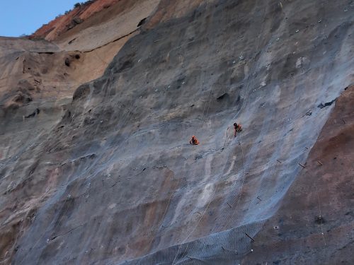 Two individuals engage in rock scaling on a cliff face. The cliff's strata display shades from light tan to deep rust, with a mesh netting partially covering the surface to catch falling debris. The workers, in bright orange safety gear, are suspended by ropes, appearing minuscule against the vastness of the cliff. Anchors and equipment are visible, dotting the surface as they systematically work across the rock, ensuring its stability. Shadows stretch over the cliff's lower portion, hinting at the setting sun.