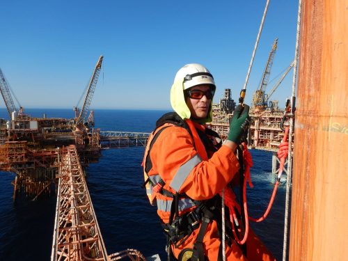 A vertech IRATA Rope Access Technician poses for a photo, suspended with North Rankin C in the background.