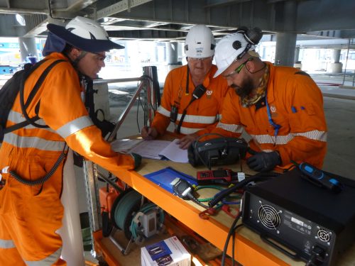 Three workers in high-visibility orange coveralls and safety helmets are examining documents and technical equipment on a portable table. They appear engaged in a serious discussion or analysis, with one writing on a paper and another looking closely at a device with dials. The environment suggests an industrial setting, with concrete structures and daylight in the background. Various equipment and tools are laid on the table, indicating an active work site.