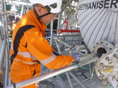 A worker wearing bright orange protective clothing and a white helmet is focused on carrying out a remote digital visual inspection (RDVI). He uses a long piece of equipment on a complex assembly of pipes and machinery surrounded by metal scaffolding. The setting suggests maintenance work in progress, highlighting the routine hands-on tasks essential for operational safety and efficiency in an industrial environment.