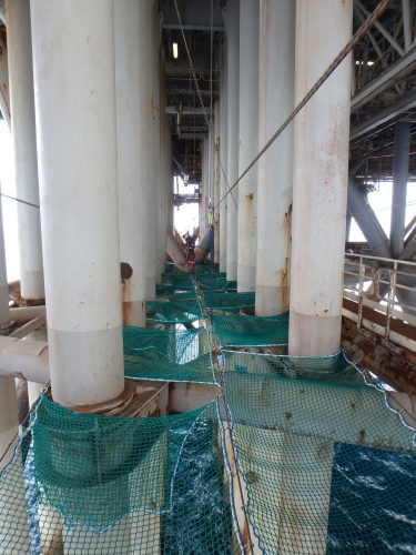 Tension netting is installed across the pillars' midway points attached to the NRA platform's underside, with the ocean below.