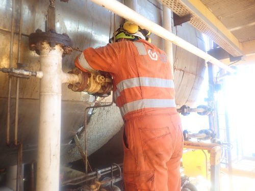 A worker wearing high-visibility orange coveralls and a safety helmet is attending to industrial piping. The worker's back is to the camera, and they appear to be examining or adjusting a section of the pipe with visible corrosion and rust. The setting is industrial, with metal structures surrounding the area and natural light from the right, indicating that the location might be an outdoor or semi-outdoor facility. Safety and maintenance work is suggested by the worker's presence and the equipment's condition.