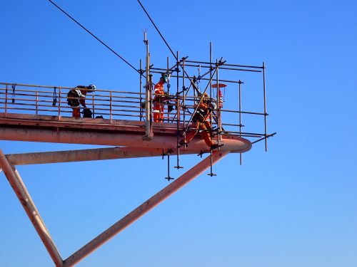 Workers assemble scaffolding on an elevated metal platform against a clear blue sky. They wear safety gear, including harnesses, helmets, and high-visibility clothing. Diagonal beams support the structure they work on and are part of an offshore facility. The workers' focused actions reflect the precision and care required for potentially hazardous construction tasks.
