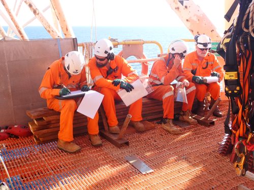 Four industrial workers in bright orange coveralls and white helmets are seated together on a deck, reviewing documents and engaged in a discussion. The sea is visible in the background, suggesting they are on an offshore platform. They are surrounded by various work equipment and safety gear, indicating a break or planning session amidst their tasks. The workers appear focused and involved in their work, highlighting the importance of teamwork and communication in such environments.