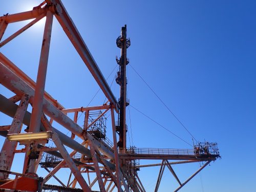 A complex metal framework against a clear blue sky, part of an industrial or offshore structure, possibly a drilling rig. The metal is painted in varying shades of white and orange, with some areas showing signs of rust and use. The horizontal and diagonal beams are visible, creating a geometric pattern with a central vertical element that may be part of the machinery. The clear sky indicates fair weather conditions for outdoor operations.