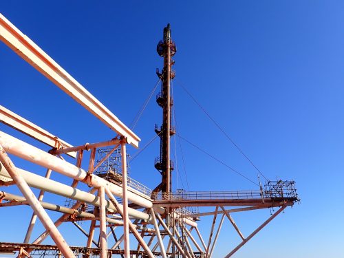 A complex metal framework against a clear blue sky, part of an industrial or offshore structure, possibly a drilling rig. The metal is painted in varying shades of white and orange, with some areas showing signs of rust and use. The horizontal and diagonal beams are visible, creating a geometric pattern with a central vertical element that may be part of the machinery. No workers are seen, but safety railings and platforms suggest that it is designed for human activity. The clear sky indicates fair weather conditions for outdoor operations.